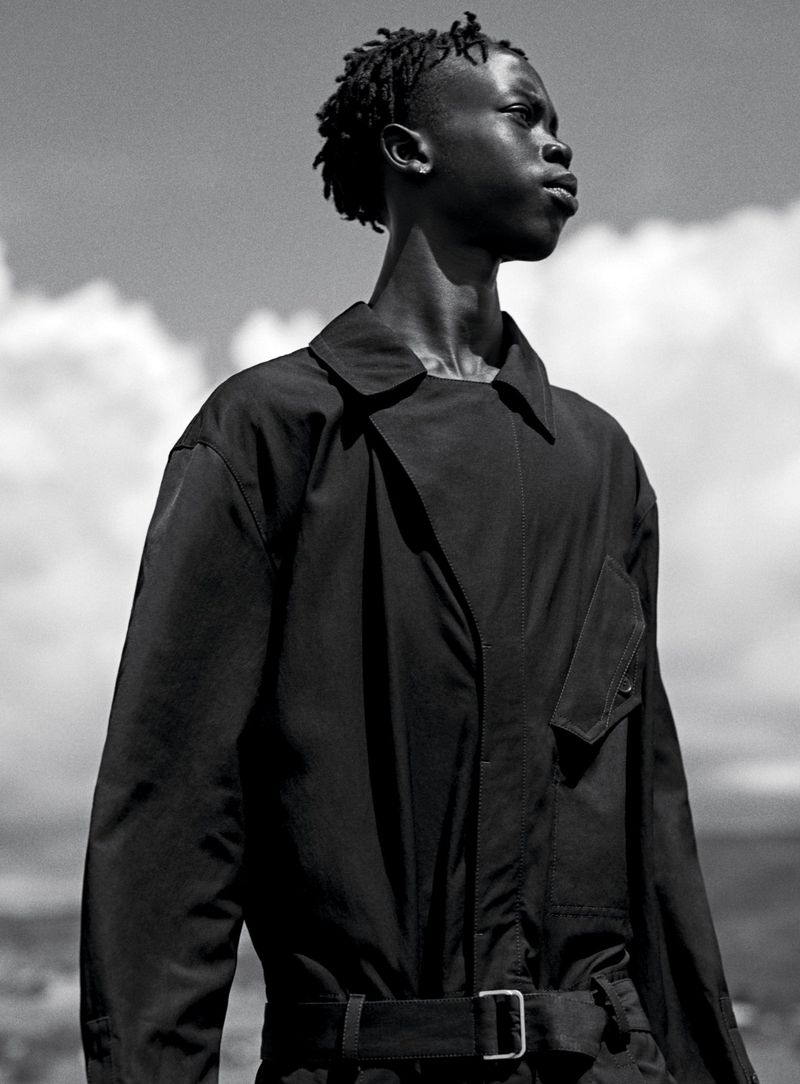Monochrome portrait of a Black male model wearing a belted black jumpsuit against a sky background with scattered clouds, looking off-camera.