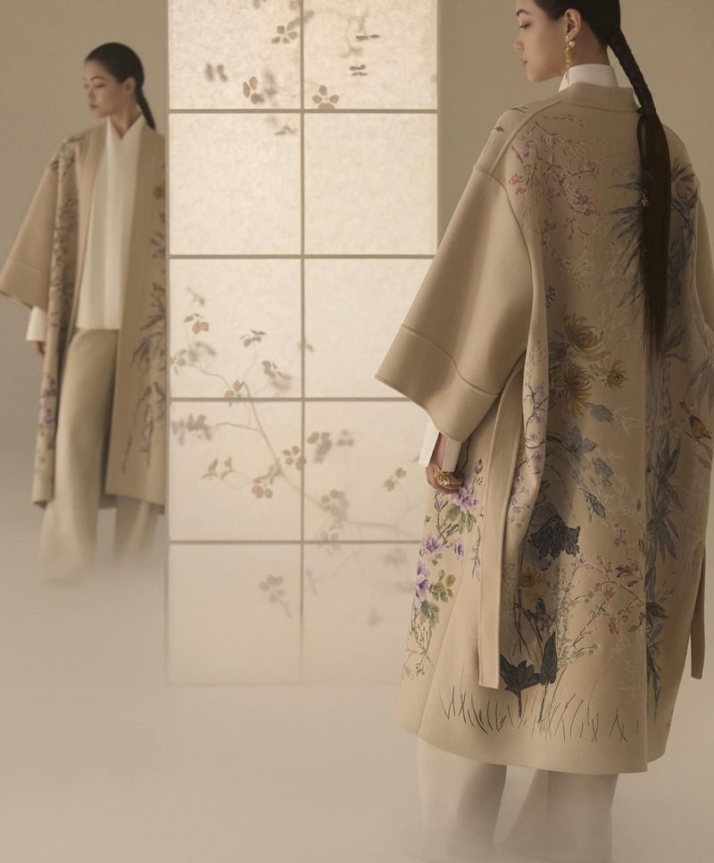 Two women in neutral-toned fashion with floral embroidery, pose in front of a paneled screen, soft lighting, and elegant styling.
