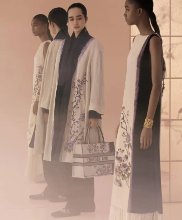 Four women in elegant dresses and a coat with floral details are posing in a studio with warm lighting, showcasing high fashion.