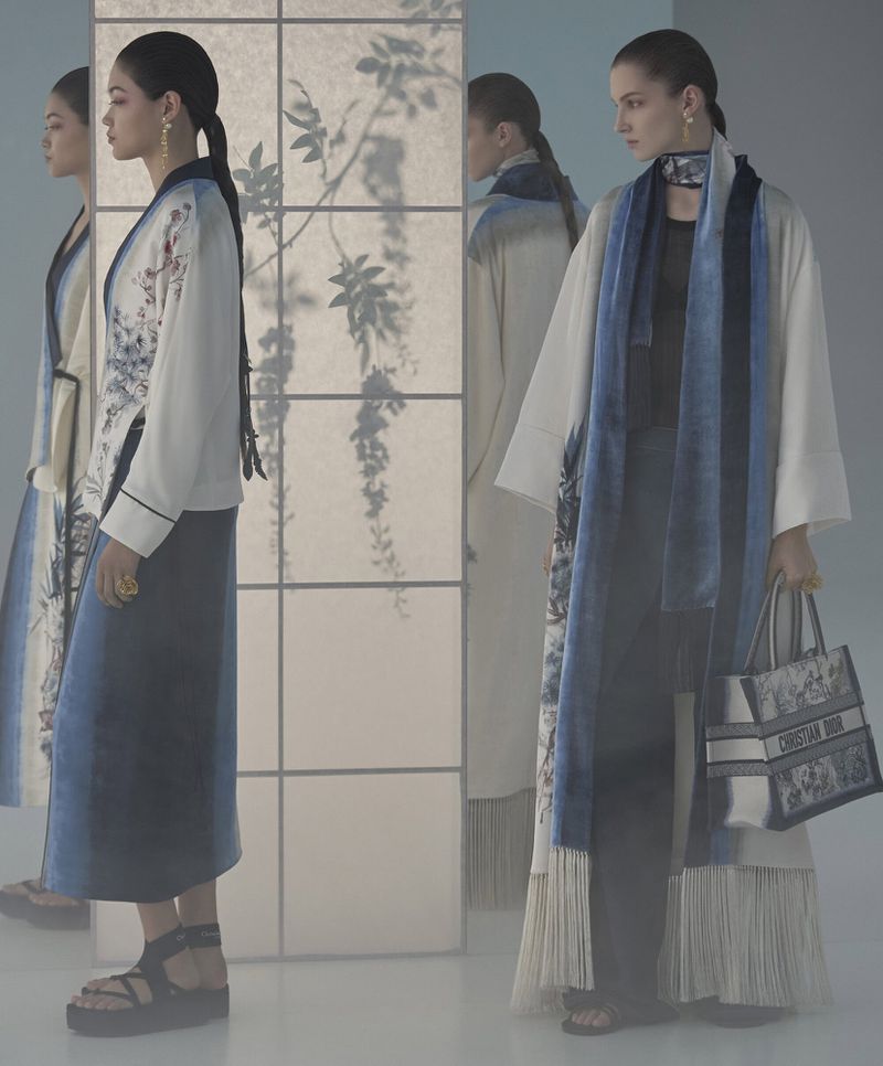 Two women wearing elegant long dresses with floral patterns and carrying a purse, posing in front of a paper screen with plant shadows.