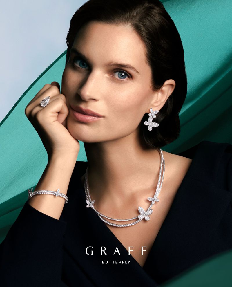 Elegant woman in black dress wearing butterfly-themed diamond jewelry set: earrings, necklace, ring, and bracelet on a teal background.