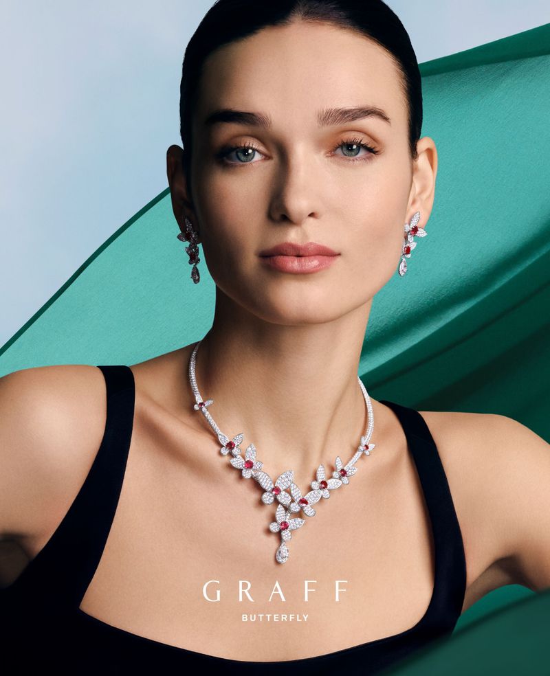 Close-up of an adult female model wearing a Graff butterfly diamond and ruby necklace and earrings against an aqua studio background.