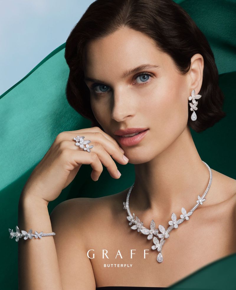 Woman presents GRAFF Butterfly jewelry collection with diamond necklace, ring, bracelet, and earrings against a draped green backdrop.