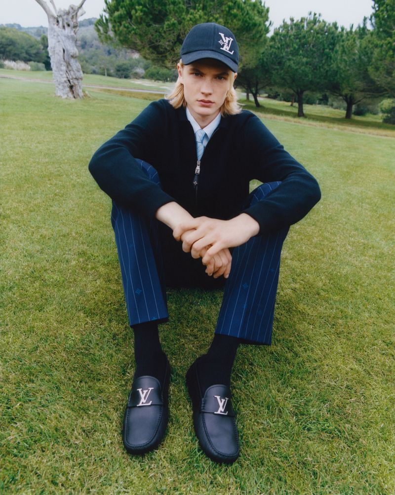 Male model wearing a cap and loafers, seated on grass outdoors, showcasing fashion and accessories, posing in natural daylight.