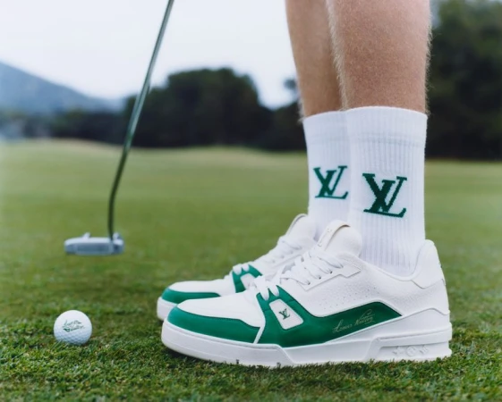 Close up of golf shoes, socks with logo, and golf ball on a green golf course with trees in the background on a cloudy day.
