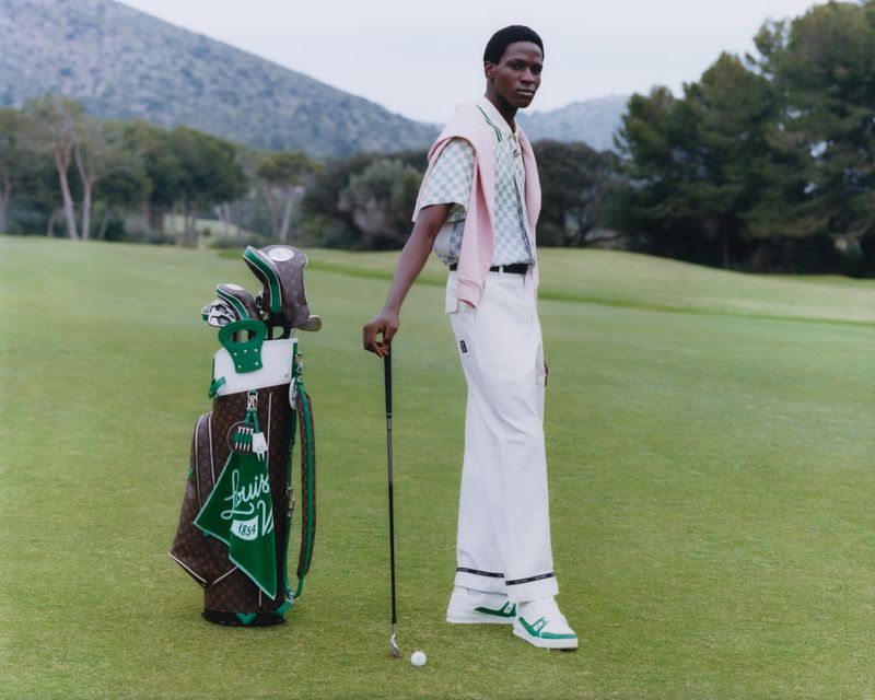 A slender Black male model poses in sportswear on a lush green golf course next to a branded golf bag, beneath a cloudy sky.