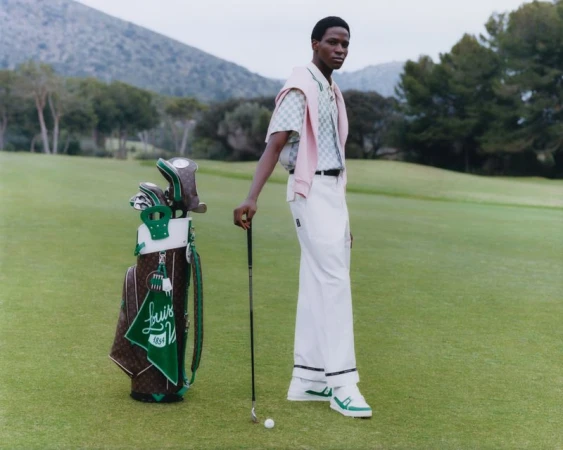 A slender Black male model poses in sportswear on a lush green golf course next to a branded golf bag, beneath a cloudy sky.