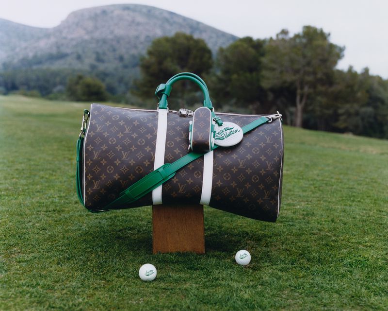 Louis Vuitton monogram duffle bag with green and white trim on a wooden block on a grassy lawn with golf balls, and mountains in background.