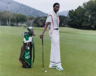 Model standing on a green golf course holding a golf club next to a monogrammed golf bag for Louis Vuitton Golf 25 Capsule