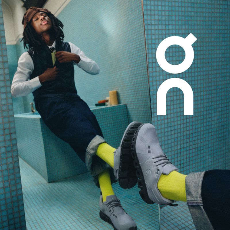 Man in vest and trousers posing with grey On shoes in a tiled bathroom setting, styled with yellow socks, and accessorized with a brown cap.