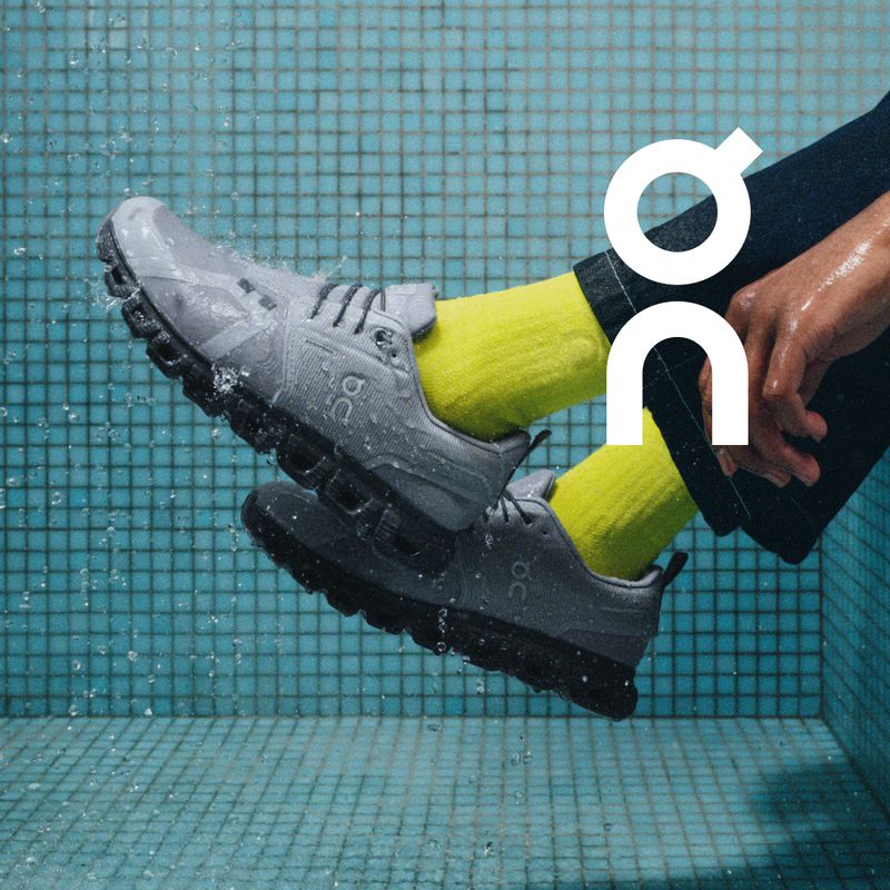 Gray running shoes by On are angled in a product shot. The shoes are being splashed with water against a blue tiled wall.