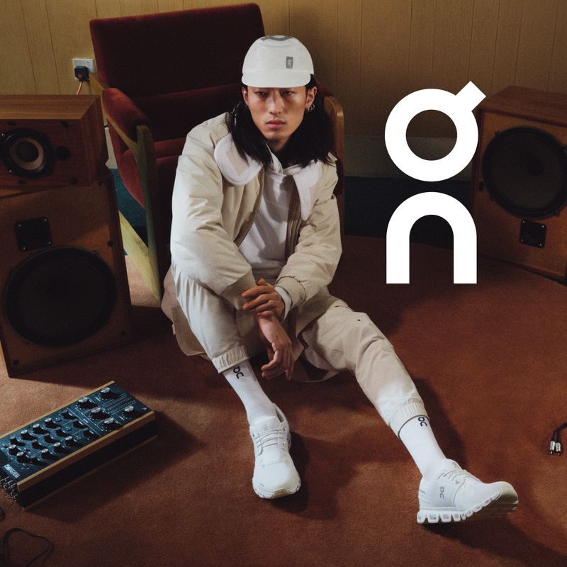 An Asian male model is seated on a brown floor wearing a white jacket, sweatpants, socks, and shoes with speakers, chair, and sound equipment.