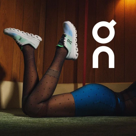 A person laying on the floor wearing blue shorts, patterned stockings, and white and blue On brand running shoes with a brown background.