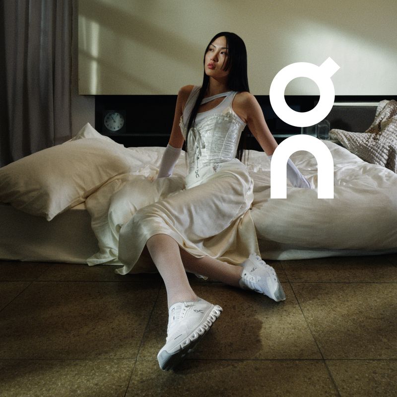 An Asian woman sits on a bed in a white corset top, dress, and athletic shoes, complemented by white opera gloves and white tights.