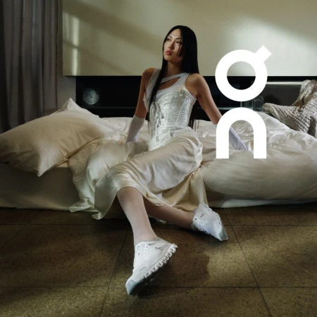 An Asian woman sits on a bed in a white corset top, dress, and athletic shoes, complemented by white opera gloves and white tights.