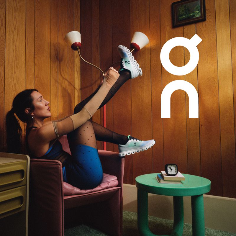 Woman in a blue dress and polka dot tights sitting in a pink chair, putting on a turquoise running shoe with the On logo in an indoor setting.