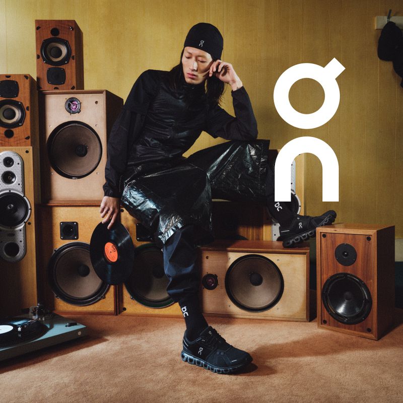 Male fashion model in a black outfit poses with vinyl record next to speakers and a record player. The On logo is visible.