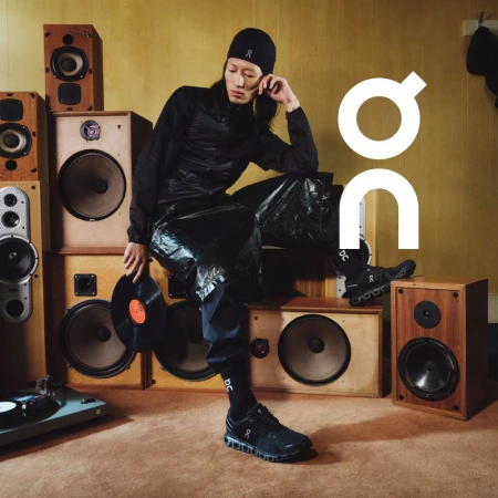 Male fashion model in a black outfit poses with vinyl record next to speakers and a record player. The On logo is visible.