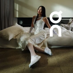 Woman in white corset and running shoes seated on a bed in a hotel room for On Running