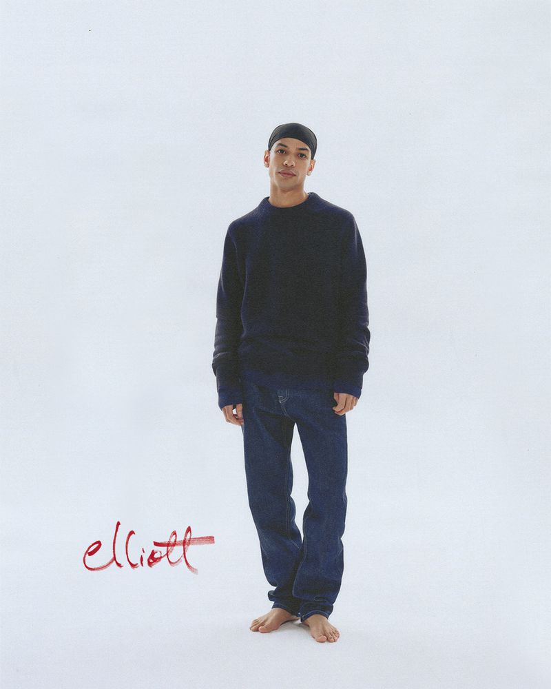 Full-length shot of an adult male in jeans and a dark sweater against a white backdrop, wearing a black beanie, with the name 'elliott' signed.