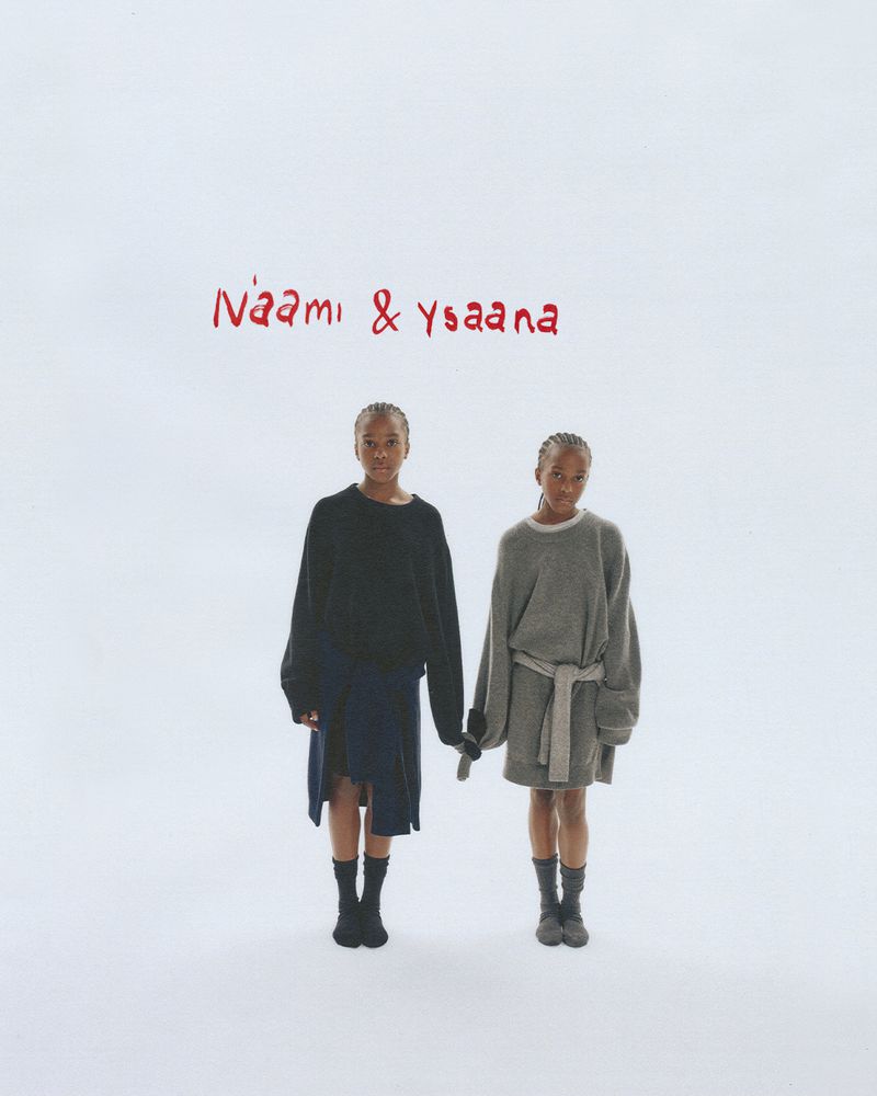 Two young, dark-skinned women stand indoors against a white background wearing matching oversized sweaters and fashionable skirts with socks.