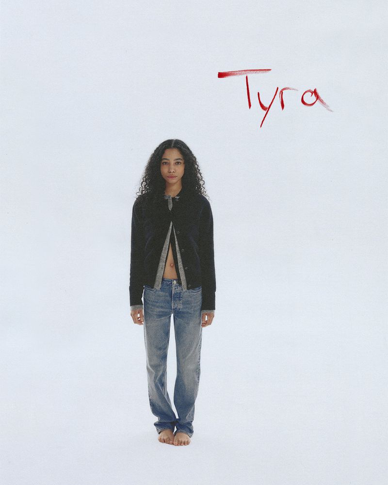 A curly-haired young woman stands barefoot wearing jeans and a black cardigan on a stark white background with 'Tyra' written in red.