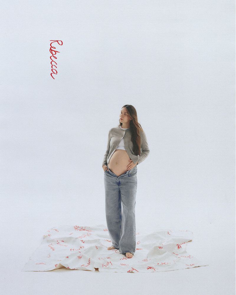 A pregnant woman in wide-leg jeans and a cardigan stands on a white sheet with red writing, set against a plain white backdrop.