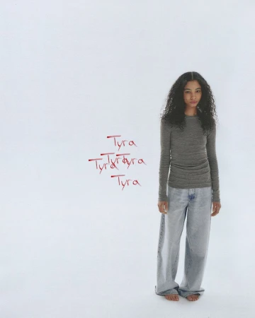 Model with long dark curly hair wearing a grey long-sleeve top and light blue jeans against a plain white studio background.