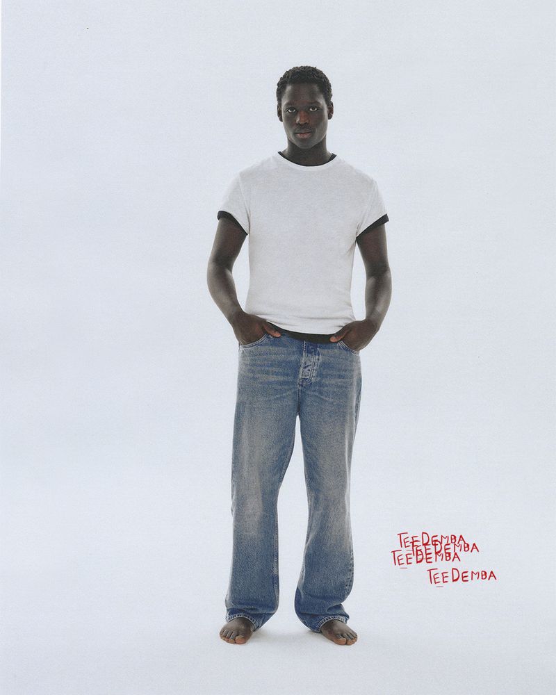 Black man wearing a white t-shirt and blue denim jeans standing against a plain light background