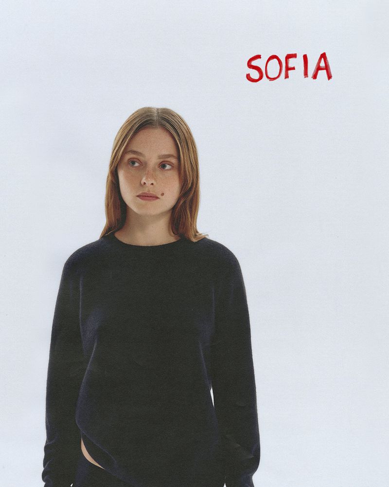 Portrait of a young woman wearing a dark crewneck sweater against a plain white background with red handwritten text.