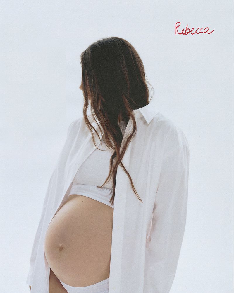 Pregnant woman in white shirt poses against bright background, showcasing baby bump and brunette hair, evoking fashion and beauty.
