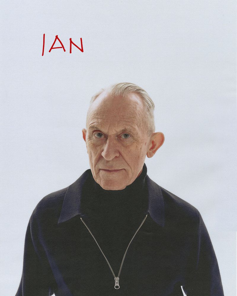 Portrait of an elderly man with short gray hair, wearing a dark blue sweater with a zipper, set against a bright white background.
