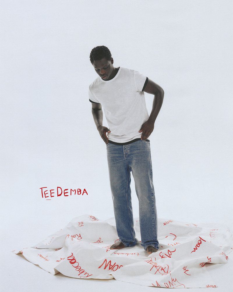 Dark-skinned male model stands on a white cloth covered in red writing, wearing a white t-shirt, blue jeans, and no shoes in a white studio.