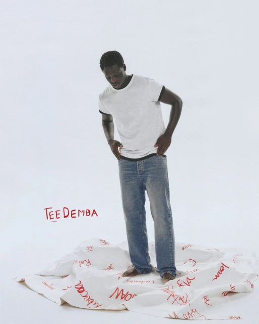 Dark-skinned male model stands on a white cloth covered in red writing, wearing a white t-shirt, blue jeans, and no shoes in a white studio.