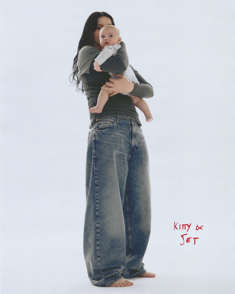 Adult woman in baggy jeans holds infant in arms in a full shot against white backdrop, featuring clear studio lighting.