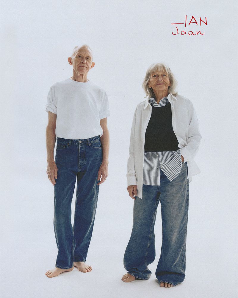 Elderly couple poses in a bright photo studio: barefoot man in jeans and t-shirt stands next to a woman in layered modern fashion.