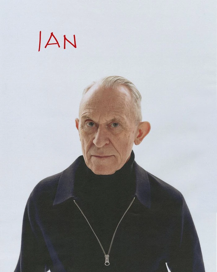 Portrait of an elderly man wearing a dark zipped sweater against a white background with red handwritten text above.