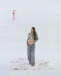 Pregnant woman wearing grey cardigan and loose blue jeans standing on a printed sheet against white background