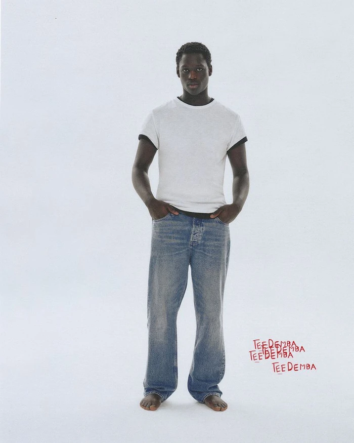 Black man wearing a white t-shirt and blue denim jeans standing against a plain light background