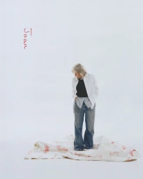 An elderly woman wearing a white shirt and denim jeans stands on a white sheet against a plain background