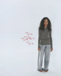 Model with long dark curly hair wearing a grey long-sleeve top and light blue jeans against a plain white studio background.