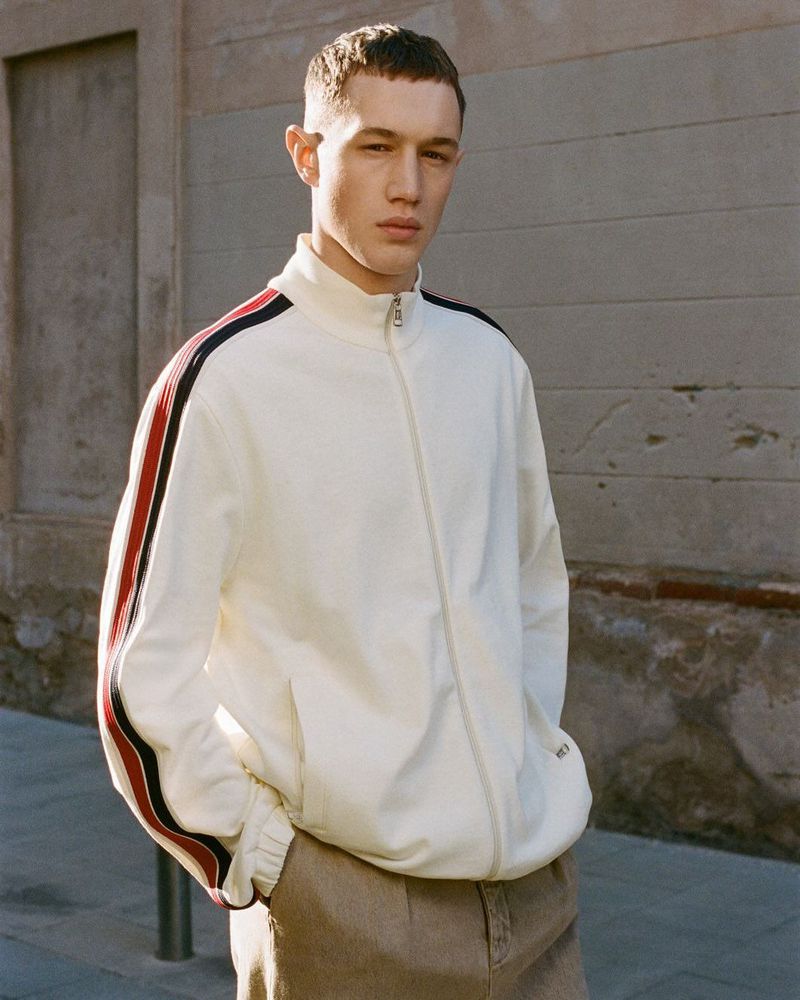 Man in white zip-up track jacket with red and black stripes standing against weathered wall outdoors in natural light.