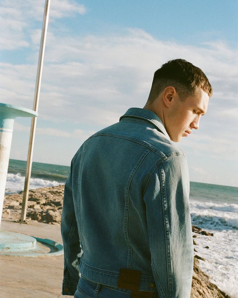 Young adult with a buzz cut, wearing a denim jacket, standing by the sea with rocks, waves, and a blue sky with scattered clouds.