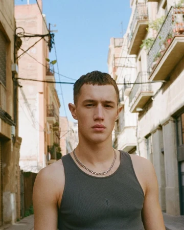 A young man in a grey tank top and silver chain stands on a narrow street with tall buildings under a clear sky in an outdoor shot.