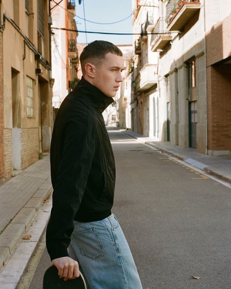 A young man wearing a black jacket and light wash jeans standing on a narrow sunny city street, holding a skateboard.