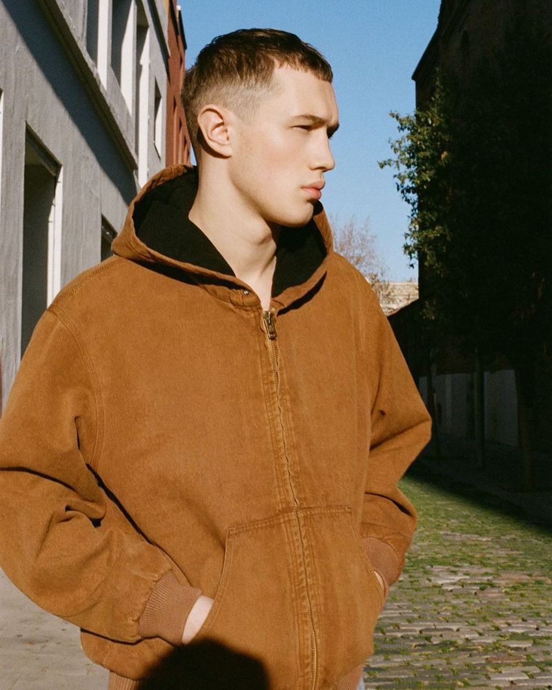 Model wearing a brown hooded canvas jacket standing on a cobblestone street lined with buildings.