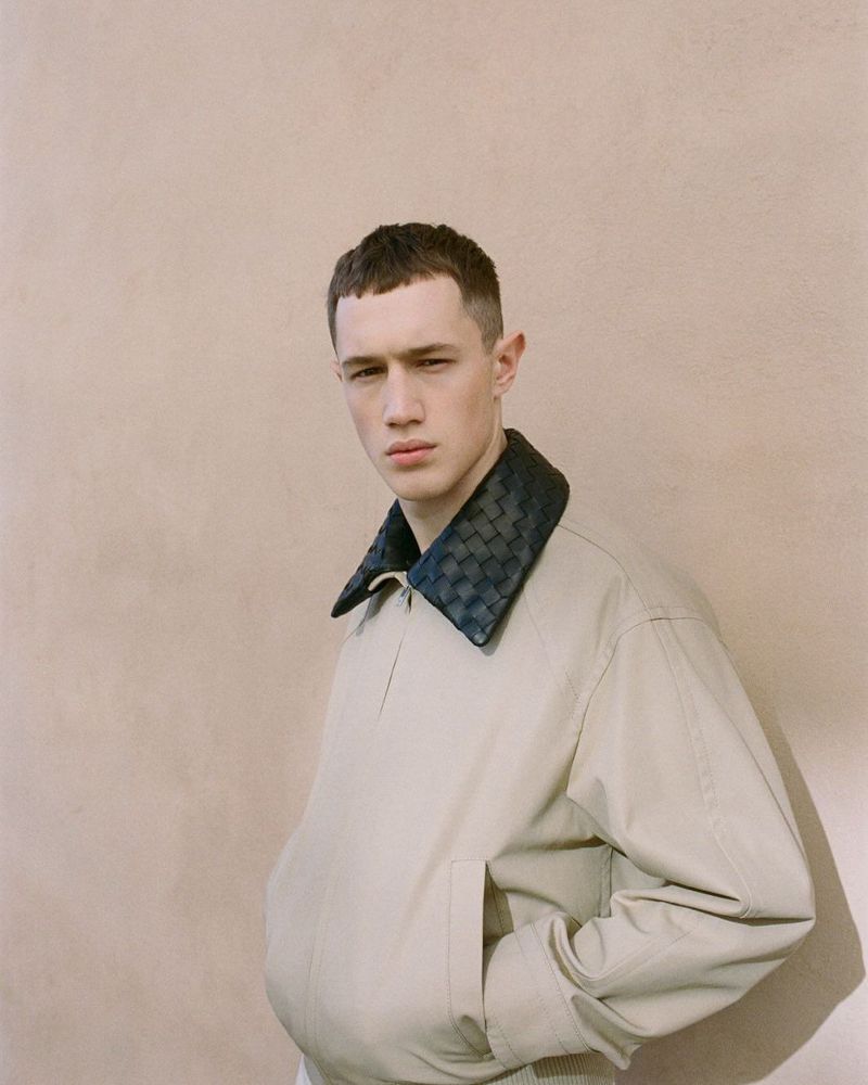 Young man wearing a cream jacket with a dark woven collar standing against a plain textured wall