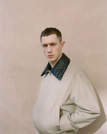 Young man wearing a cream jacket with a dark woven collar standing against a plain textured wall