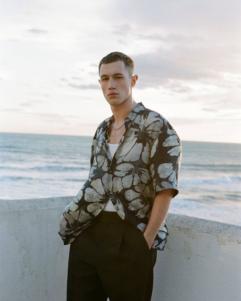 A stylish young man in a floral shirt poses confidently outdoors against a beach backdrop, showcasing a blend of casual and contemporary fashion.