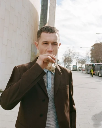 Man in a brown blazer holds a paper bag to his mouth on a city sidewalk, with blurred bus in the background on a sunny day.
